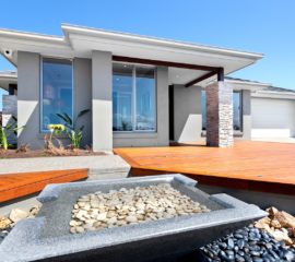 Well decorated yard and garden using stone elements in front of a modern house, closeup of a small square shape pond made by pure stone included gravel and water inside of it. There are white and black rocks around the pond. The wooden yard is included rectangle shape bars, a garage can be seen from the right side. Inside of the mansion is seen through the window